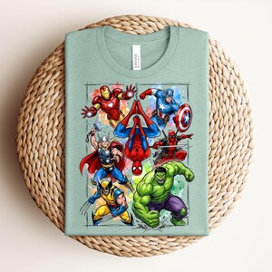 May include: A pale green t-shirt with a vibrant graphic of superheroes such as Iron Man, Captain America, and Spider-Man. The design is framed with a watercolour-style border. The shirt is folded on a woven surface.