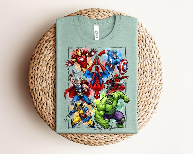 May include: A pale green t-shirt with a vibrant graphic of superheroes such as Iron Man, Captain America, and Spider-Man. The design is framed with a watercolour-style border. The shirt is folded on a woven surface.