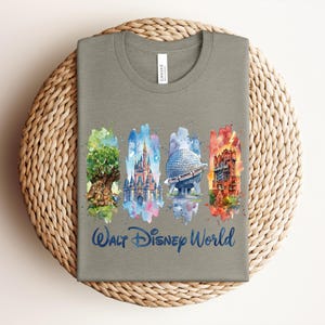 May include: A gray t-shirt featuring watercolor illustrations of iconic Walt Disney World landmarks: the Tree of Life, Cinderella Castle, Spaceship Earth, and the Tower of Terror. The words "Walt Disney World" are written in a blue script font below the illustrations.
