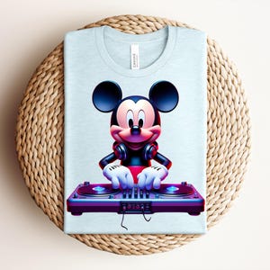 May include: Light blue t-shirt featuring a colorful graphic of Mickey Mouse as a DJ. Mickey is wearing headphones and has his hands on a purple and blue turntable. The shirt is folded on a woven, circular surface.