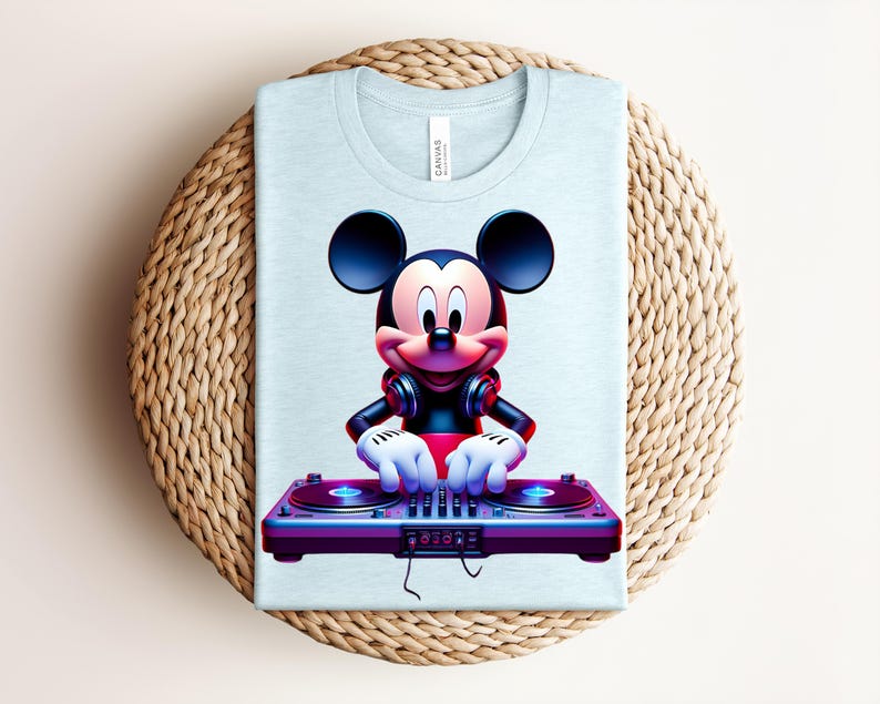 May include: Light blue t-shirt featuring a colorful graphic of Mickey Mouse as a DJ. Mickey is wearing headphones and has his hands on a purple and blue turntable. The shirt is folded on a woven, circular surface.