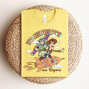 May include: A yellow t-shirt featuring a graphic print of Buzz Lightyear and Woody riding a rocket ship. The text "TO INFINITY" is arched above the characters, and "AND BEYOND" is printed below the rocket, surrounded by small clouds and stars.