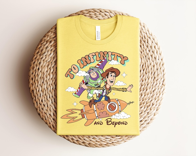 May include: A yellow t-shirt featuring a graphic print of Buzz Lightyear and Woody riding a rocket ship. The text "TO INFINITY" is arched above the characters, and "AND BEYOND" is printed below the rocket, surrounded by small clouds and stars.