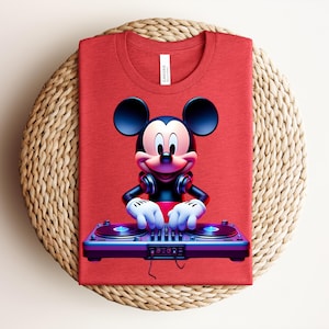 May include: A red t-shirt featuring a graphic of Mickey Mouse as a DJ, wearing headphones and operating a turntable. The shirt is folded and placed on a woven, circular mat. The tag reads "Canvas".