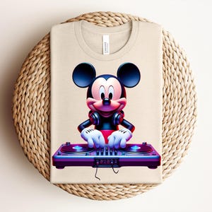 May include: Beige t-shirt featuring a vibrant graphic of Mickey Mouse as a DJ. Mickey is wearing headphones and has his hands on a purple and blue DJ mixer. The shirt is folded on a woven, circular surface.