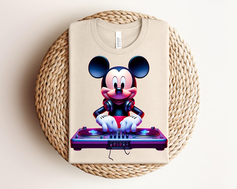 May include: Beige t-shirt featuring a vibrant graphic of Mickey Mouse as a DJ. Mickey is wearing headphones and has his hands on a purple and blue DJ mixer. The shirt is folded on a woven, circular surface.