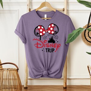 May include: A lilac-colored t-shirt featuring a graphic design for a Disney trip. The design includes a red and white polka-dot bow, Mickey Mouse ears, a castle silhouette, and the words "Disney Trip" in red.