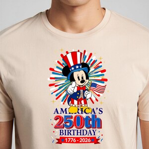 May include: Beige t-shirt featuring a graphic of Mickey Mouse dressed as Uncle Sam, holding an American flag. The design celebrates America's 250th birthday, with text reading "AMERICA'S 250th BIRTHDAY 1776-2026".