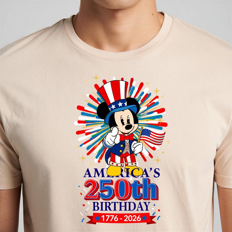 May include: Beige t-shirt featuring a graphic of Mickey Mouse dressed as Uncle Sam, holding an American flag. The design celebrates America's 250th birthday, with text reading "AMERICA'S 250th BIRTHDAY 1776-2026".