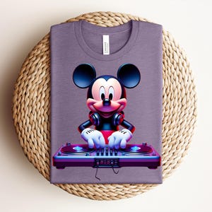 May include: A purple t-shirt featuring a vibrant graphic of Mickey Mouse as a DJ. Mickey is wearing headphones and has his hands on a turntable. The shirt is folded and placed on a woven, circular mat.