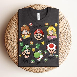 May include: Dark gray t-shirt featuring colorful illustrations of Super Mario characters, including Mario, Luigi, Princess Peach, Daisy, Yoshi, and Toad. The words "Super Mario" and character names are also printed on the shirt.
