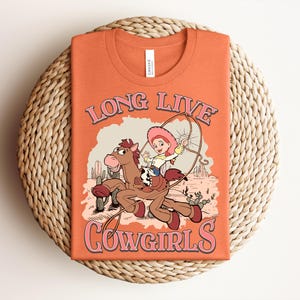 May include: An orange t-shirt featuring a graphic of a cartoon cowgirl riding a brown horse in a desert landscape with cacti. The text "LONG LIVE COWGIRLS" is printed in pink with a dark outline above and below the illustration of the character.