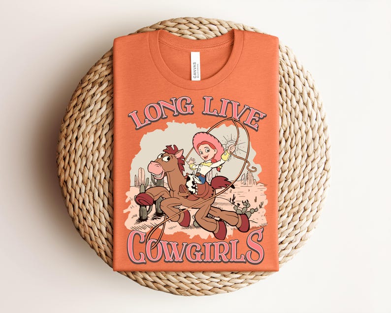 May include: An orange t-shirt featuring a graphic of a cartoon cowgirl riding a brown horse in a desert landscape with cacti. The text "LONG LIVE COWGIRLS" is printed in pink with a dark outline above and below the illustration of the character.