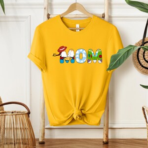 May include: Yellow t-shirt with the word "MOM" in colorful, cartoon-style letters. The "O" is designed with a cloud pattern, and the shirt is knotted at the bottom.