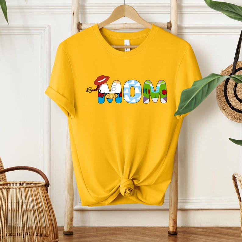 May include: Yellow t-shirt with the word "MOM" in colorful, cartoon-style letters. The "O" is designed with a cloud pattern, and the shirt is knotted at the bottom.