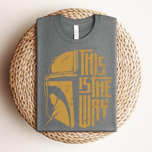 May include: A dark gray crew neck t-shirt featuring a distressed gold graphic of a helmet silhouette and the text THIS IS THE WAY. The design is centered on the chest of the shirt, which has a standard short-sleeve cut and a visible brand label.