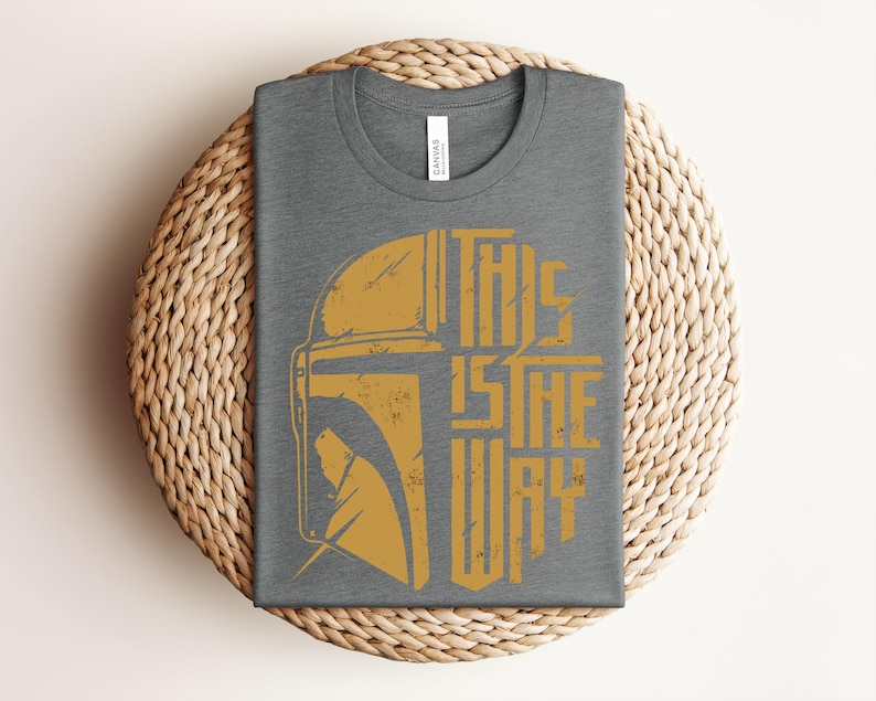 May include: A dark gray crew neck t-shirt featuring a distressed gold graphic of a helmet silhouette and the text THIS IS THE WAY. The design is centered on the chest of the shirt, which has a standard short-sleeve cut and a visible brand label.