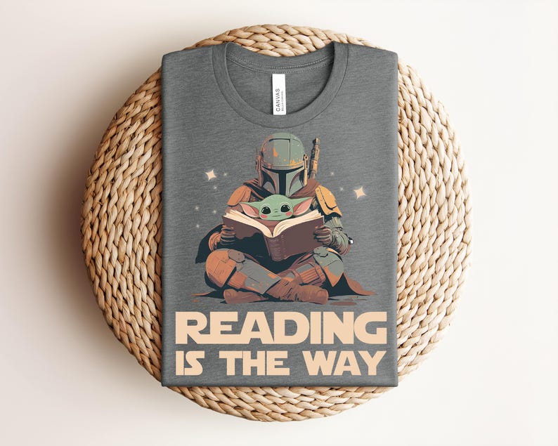 May include: A heather gray t-shirt featuring a graphic of a helmeted character sitting cross-legged and reading a book to a small green creature. The text below the illustration reads READING IS THE WAY in large, bold, light-colored capital letters.