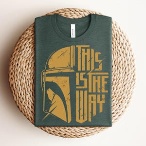 May include: A dark green crew neck t-shirt featuring a distressed gold graphic of a helmet and the text THIS IS THE WAY. The shirt has a white brand label at the inner neckline and is displayed folded to showcase the front design clearly.