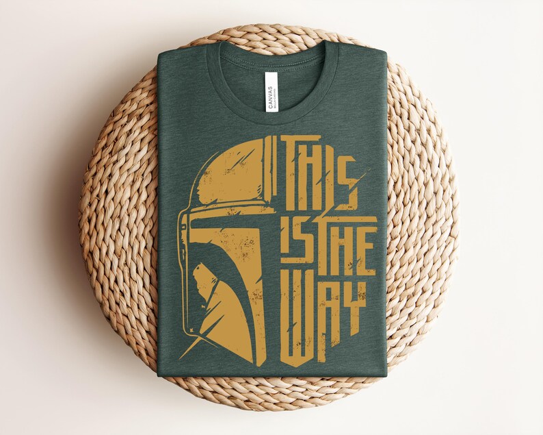 May include: A dark green crew neck t-shirt featuring a distressed gold graphic of a helmet and the text THIS IS THE WAY. The shirt has a white brand label at the inner neckline and is displayed folded to showcase the front design clearly.