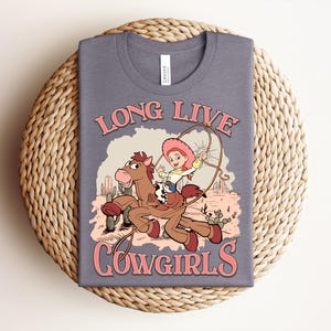 May include: A heather grey t-shirt featuring a graphic print of a cartoon character riding a brown horse in a desert landscape with cacti. The text "LONG LIVE COWGIRLS" is printed in pink above and below the illustration, which includes a lasso.