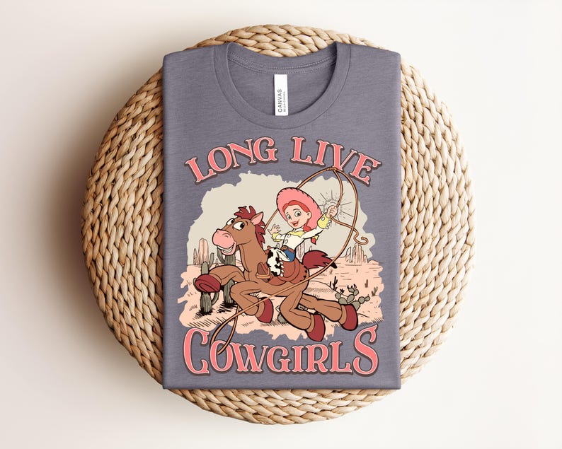 May include: A heather grey t-shirt featuring a graphic print of a cartoon character riding a brown horse in a desert landscape with cacti. The text "LONG LIVE COWGIRLS" is printed in pink above and below the illustration, which includes a lasso.