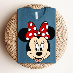 May include: A blue t-shirt featuring a graphic of Minnie Mouse's face. The design includes a red bow with white polka dots, black ears, and a smiling face. The shirt is folded and placed on a woven surface.