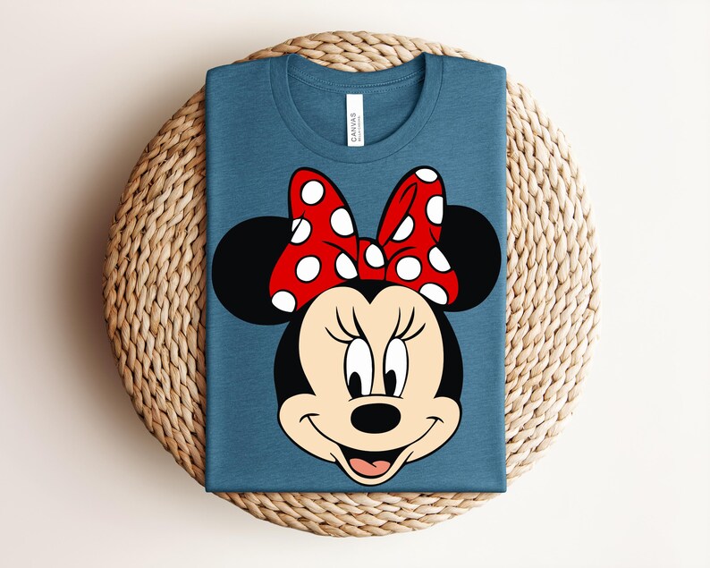 May include: A blue t-shirt featuring a graphic of Minnie Mouse's face. The design includes a red bow with white polka dots, black ears, and a smiling face. The shirt is folded and placed on a woven surface.