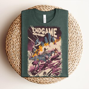 Pu&ograve; includere: Una t-shirt verde bosco scuro con una stampa grafica che riporta il testo ENDGAME in alto. L'illustrazione raffigura una scena di battaglia con figure stilizzate, terreno roccioso, fiamme arancioni e fulmini viola su sfondo scuro.