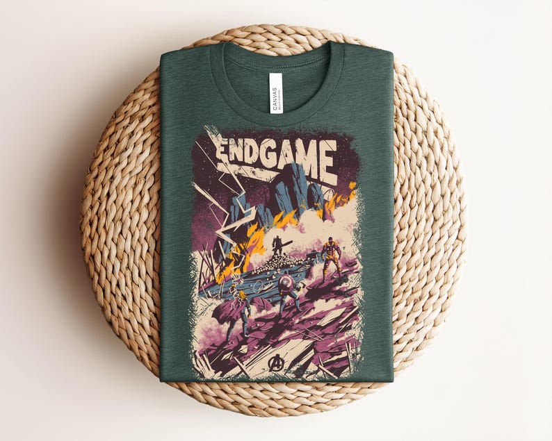 Pu&ograve; includere: Una t-shirt verde bosco scuro con una stampa grafica che riporta il testo ENDGAME in alto. L'illustrazione raffigura una scena di battaglia con figure stilizzate, terreno roccioso, fiamme arancioni e fulmini viola su sfondo scuro.