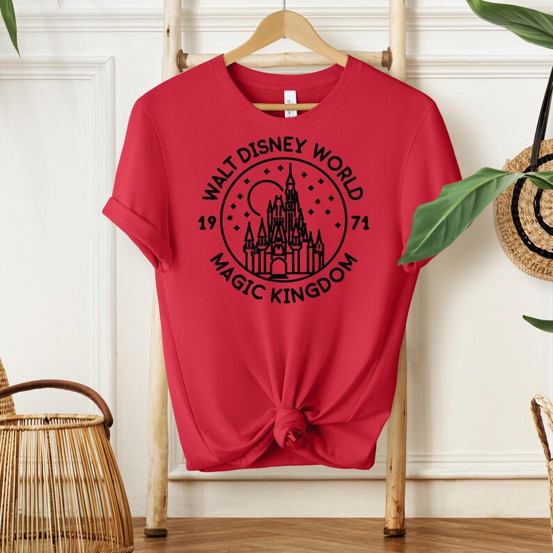 May include: A red t-shirt with a black graphic of the Walt Disney World logo, featuring a castle, moon, and stars. The text reads "Walt Disney World Magic Kingdom 1971". The shirt is knotted at the waist and hangs on a wooden hanger.