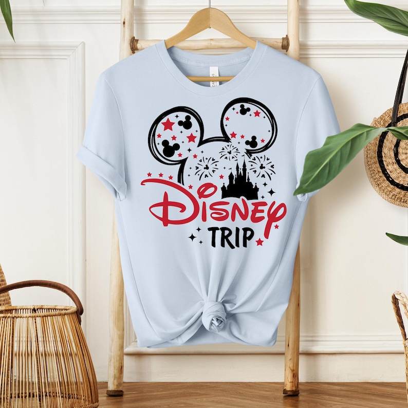 May include: Light blue t-shirt featuring a graphic design with Mickey Mouse ears, stars, fireworks, and a castle silhouette. The text "Disney Trip" is in red. The shirt is displayed on a wooden hanger.
