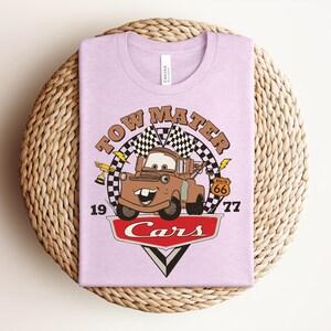 May include: A light purple t-shirt featuring a graphic of the character Tow Mater from the movie Cars. The design includes a checkered flag, a gold trophy, a Route 66 sign, the text Tow Mater, 1977, and the Cars logo in red and white.