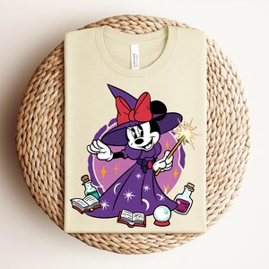 May include: A light beige t-shirt featuring a cartoon Minnie Mouse dressed as a witch. She wears a purple dress, a witch hat with a red bow, and holds a wand. The design includes potion bottles, a crystal ball, and open books.