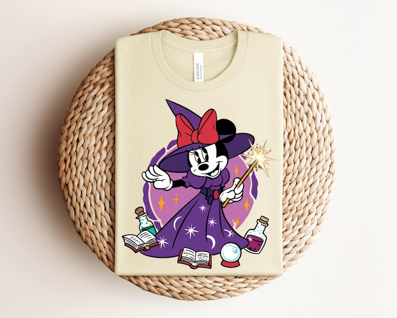 May include: A light beige t-shirt featuring a cartoon Minnie Mouse dressed as a witch. She wears a purple dress, a witch hat with a red bow, and holds a wand. The design includes potion bottles, a crystal ball, and open books.