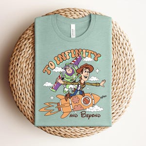 May include: A light green t-shirt featuring a graphic of Buzz Lightyear and Woody from Toy Story. Woody rides an orange rocket while Buzz flies alongside. The text reads TO INFINITY AND BEYOND in a retro font, surrounded by small clouds and stars.