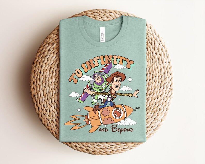 May include: A light green t-shirt featuring a graphic of Buzz Lightyear and Woody from Toy Story. Woody rides an orange rocket while Buzz flies alongside. The text reads TO INFINITY AND BEYOND in a retro font, surrounded by small clouds and stars.