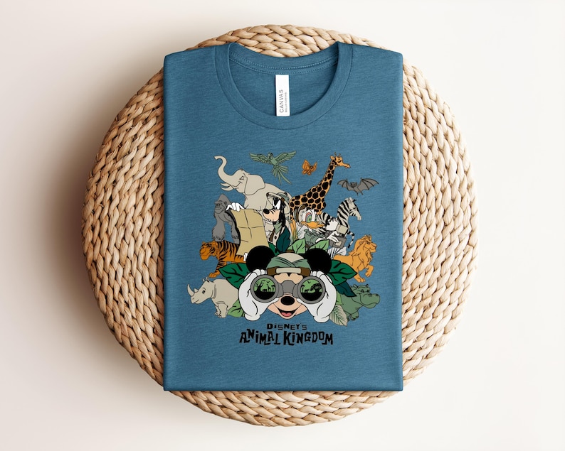 Puede incluir: Una camiseta azul con un estampado gr&aacute;fico de Disney's Animal Kingdom que muestra a Mickey Mouse mirando a trav&eacute;s de binoculares, rodeado de animales como un elefante, jirafa, tigre, cebra y rinoceronte, con el texto Disney's Animal Kingdom.