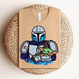 Puede incluir: Una camiseta beige con una ilustraci&oacute;n de dibujos animados de un personaje mandaloriano con armadura, sosteniendo a un alien&iacute;gena verde. El dise&ntilde;o incluye detalles azules y plateados, sobre un fondo espacial. La camiseta est&aacute; doblada sobre una superficie tejida.