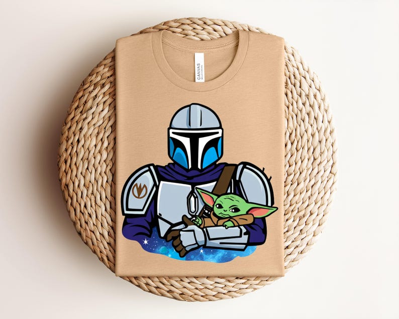 Puede incluir: Una camiseta beige con una ilustraci&oacute;n de dibujos animados de un personaje mandaloriano con armadura, sosteniendo a un alien&iacute;gena verde. El dise&ntilde;o incluye detalles azules y plateados, sobre un fondo espacial. La camiseta est&aacute; doblada sobre una superficie tejida.