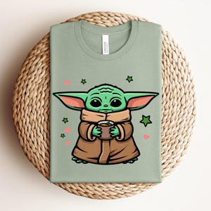 May include: Sage green t-shirt featuring a cartoon illustration of a green alien character holding a cup. The character is wearing a brown cloak and has large ears. Pink hearts and green stars decorate the shirt.