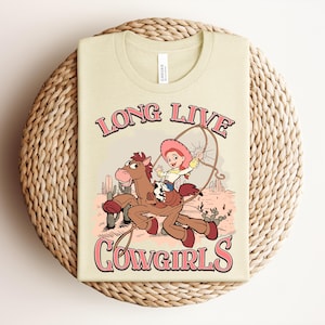 May include: A cream-colored t-shirt featuring a graphic of a cowgirl riding a brown horse in a desert landscape. The text "LONG LIVE COWGIRLS" is printed in pink with a dark outline above and below the illustration, which includes cacti.