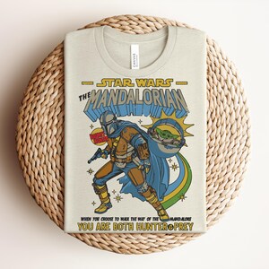 Puede incluir: Una camiseta de color crema con un gr&aacute;fico de The Mandalorian y Grogu. El texto dice Star Wars The Mandalorian, Bounty Hunter y When you choose to walk the way of the Mandalore you are both hunter and prey en amarillo y azul.