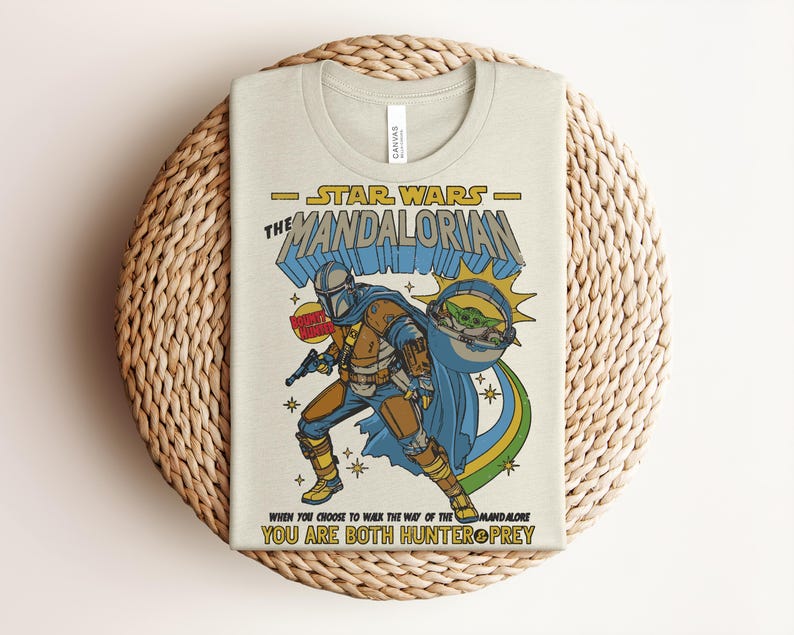 Puede incluir: Una camiseta de color crema con un gr&aacute;fico de The Mandalorian y Grogu. El texto dice Star Wars The Mandalorian, Bounty Hunter y When you choose to walk the way of the Mandalore you are both hunter and prey en amarillo y azul.