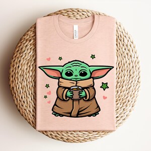 May include: A light pink t-shirt featuring a cartoon character with large green ears, holding a cup. The character is wearing a brown coat and a tan scarf. Green stars and pink hearts are scattered around the character.