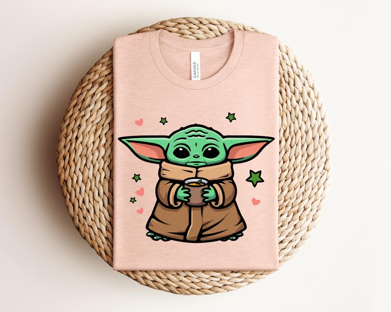 May include: A light pink t-shirt featuring a cartoon character with large green ears, holding a cup. The character is wearing a brown coat and a tan scarf. Green stars and pink hearts are scattered around the character.