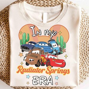 K&ouml;nnte beinhalten: Cremefarbenes T-Shirt mit einer Grafik von Zeichentrickautos aus dem Film Cars. Das Design enth&auml;lt den Text "In my Radiator Springs ERA" mit Kakteen und Sternen. Das Shirt hat einen Rundhalsausschnitt.