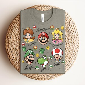 May include: A gray t-shirt featuring Super Mario characters: Mario, Luigi, Princess Peach, Daisy, Yoshi, and Toad. The shirt includes the words "Super Mario", "Daisy", "Luigi", "Princess Peach", "Yoshi", and "Toad". The design includes stars, coins, and mushrooms.