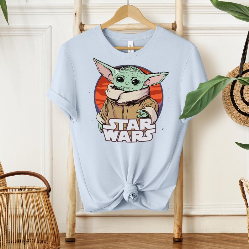 May include: Light blue t-shirt featuring a graphic of a green alien character from Star Wars. The character is set against a red and orange background, with the words "STAR WARS" below. The shirt is hanging on a wooden hanger.