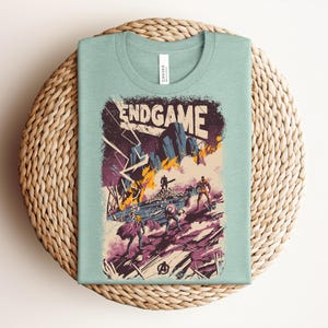 Pu&ograve; includere: Una t-shirt girocollo verde chiaro con una stampa grafica che riporta il testo ENDGAME in alto. L'illustrazione raffigura una scena di battaglia con diverse figure, fulmini, fuoco e detriti nei toni del viola, arancione e blu scuro.